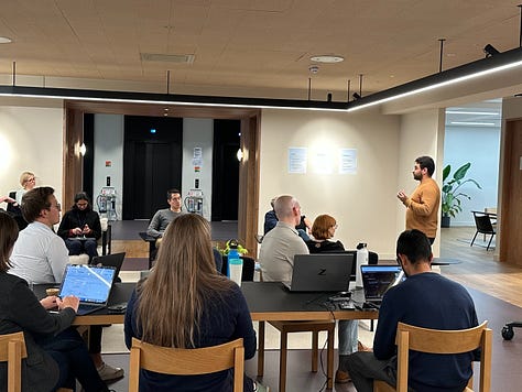 1. A photo of a sign saying 'Cambridge Neuroworks, Powered by ARIA, Cambridge and the Advanced Research + Invention Agency (ARIA). Accelerating the translation of ideas to find revolutionary neurotechnologies. There are people mingling in the background of the image. 3. A photo of a person presenting to a room full of people, there is a screen in the background saying 'Powered by ARIA' and the Fifty Years logo on it. 4. A photo of people mingling at an event. In the background there is a large vinyl decal saying 'Hello brain!' 5. A room full of people listening to someone speak (off camera) at a Pillar VC event. 6. A person presenting to a room of people. 