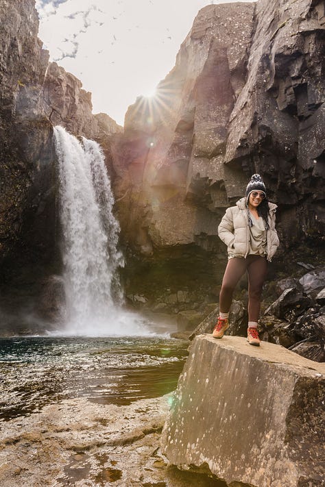 Photo of woman smiling over her shoulder sitting atop a boulder and wearing winter clothes, photo of a white and red church atop a green and yellow hill, photo of a woman wearing winter clothes smiling sitting on a boulder along a glacial lagoon river with large chunks of ice, photo of a small tow along a green coast with a jagged green and brown mountain rising up from the coastal edge in the background, photo of a woman standing on a boulder with a waterfall in the background, photo of a jagged green and gray rocky canyon with a river winding through, photo of large chunks of glacial ice on a beach, photo of two men and two women staggered on a rainbow-colored road that leads to a church, photo of the interior of a diner with a red cooler and a woman serving coffee