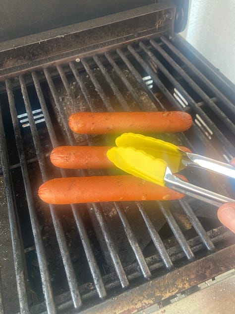 Grilled hotdogs being prepared on an outdoor grill with a number of ingredients and the buns.