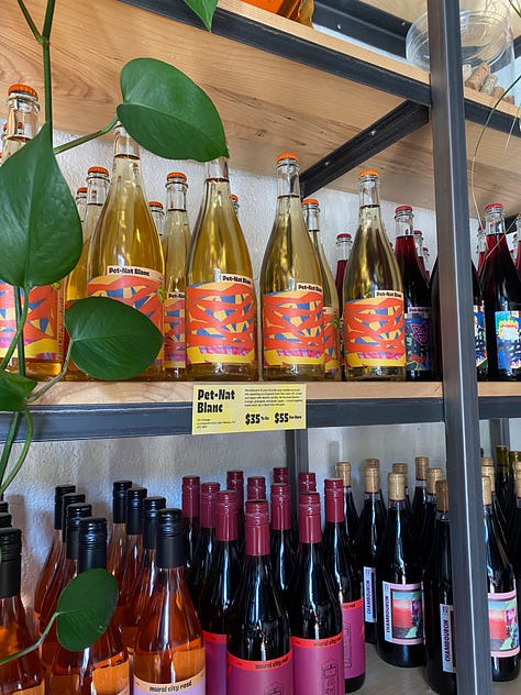 Wine bottles with images of my artwork on the labels arranged in rows on shelves. In the middle is a photo of me, a man holding a bottle of the Pet-Nat Rouge smiling for the photo and looking out at the viewer.