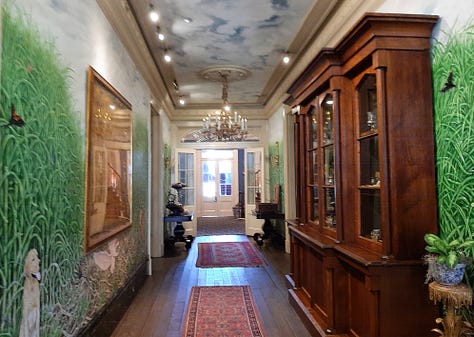 The interior of Houmas House is stately and beautiful with a wide center hall with large portraits on the wall. The second photo shows a fireplace and artwork; the third photo shows a desk in a stately room with a portrait in an ornate frame. The last photos are of bedrooms and a curving staircase.