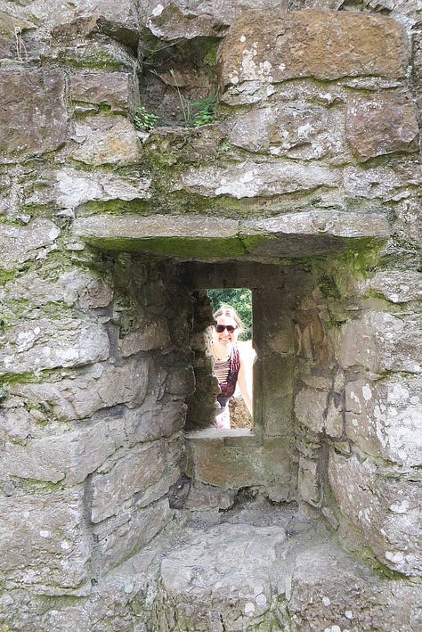 A woman in a red and black dress, bending down to look into a drainage hole in a wall in Pendragon Castle, a 12th-century castle in Cumbria, England; A woman wearing sunglasses, peering through a medieval squint at Penmon Dovecote, built c. 1600, near Beaumaris in North Wales; a woman wearing a red t-shirt, black trousers and a hat standing in a walled street in Split, Croatia. 