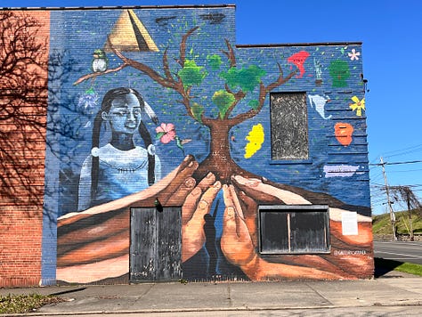 A mural on a brick building. The image of large hands supporting a tree reaching up to the sky, and a girl with pigtails standing in the near background.