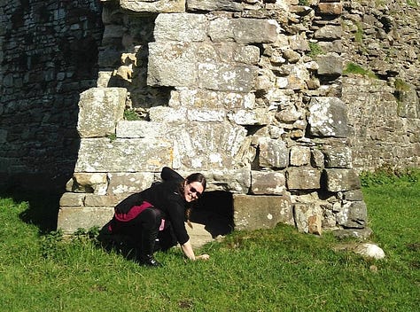 A woman in a red and black dress, bending down to look into a drainage hole in a wall in Pendragon Castle, a 12th-century castle in Cumbria, England; A woman wearing sunglasses, peering through a medieval squint at Penmon Dovecote, built c. 1600, near Beaumaris in North Wales; a woman wearing a red t-shirt, black trousers and a hat standing in a walled street in Split, Croatia. 