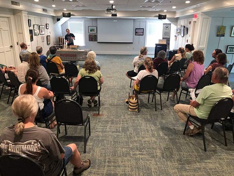 In the first photo James stands behind a table with a blanket printed witht he words "Mushroom Microfarm". Second picture, James and table are in the background with class attendees at forefront. Third picture, close up of tinctures on the table.