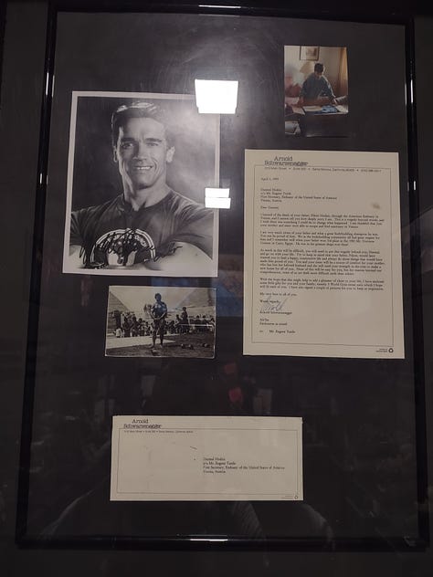 The top left image shows a letter from Arnold Schwarzenegger to the zon of Yugoslavian bodybuilding champion, Fikret Hodzic. The top middle image shows the gramed letter together with a picture of Schwarzenegger . The top right image shows a bodybuilding shirt from Schwarzenegger in yellow that says "World Gym." The bottom left image shows the text describing Fikret Hodzic's life and death. The bottom right image shows a gold medal won by Fikret Hodzic at the European Bodybuilding Championships.