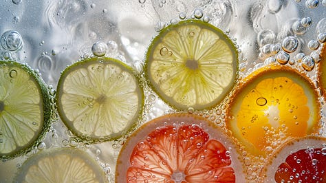  An arrangement of various citrus slices – lemon, lime, orange, and grapefruit – submerged in sparkling water, with bubbles clinging to the fruit, set against a light box for a luminous effect