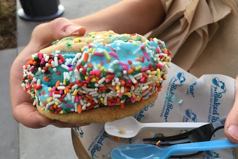 Three images of large delightful cookies with blue ice cream in the middle and toffee chunks or sprinkles around the outside edges of the ice cream, all being held in wrappers that say The Baked Bear.