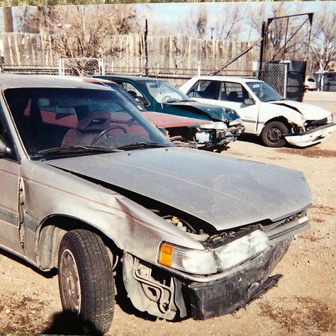 My champagne Mazda 626 lurches cockeyed in the wreck yard with other munched vehicles. The smashed front bumper lies on the ground. The windshield is lanced with cracks. The passenger side is caved in. The drivers side is demolished.