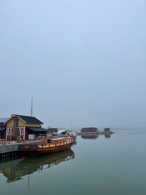 A blue sky with clouds and sky reflected in the water, a foggy day with grey sky, grey, still water and a half built wooden boat in the water moored to the pier, 3: looking out to water and fog with wires from rigging spanning the photo corners and the end gear of the deck railing in the foreground. 