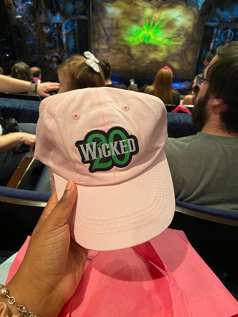   From top left to bottom right - going across horizontally. Image 1) Alexa is holding up a green frisbee that says Defying Gravity since 2003 in black font. The text is curved around the frisbee, in a half circle. In the center, there is a black stenciled picture of Elphaba holding up her Broome. In the middle of skirt, the text “Wicked” is printed with a 20 in the background of the word. Both ‘Wicked’ and the number ’20’ are highlighted in green font. Image 2) The back of Alexa’s left hand. She has a temporary tattoo printed that reads “Toss Toss” in red cursive ink. The two T’s are connected, and there is an asterix/flower type symbol over the second to last S. In smaller font, “Wicked” is printed in all caps. Image 3) Alexa is posing in front of a replica of the clocktower, as seen in the Wicked production. At the top of the clock tower, which is wooden, with antique looking wheels and roman numerals, there is a dragon neck (with its head cut off in the photo) with webbed looking wings. Alexa posing side wise, with a surprised expression on her face, as if she is being scared by someone. She is wearing a polka dotted scarf in her hair, and a pink collared shirt (you can only see her from the shoulders up.) Image 4) Alexa is posing with Carrie St. Louis, who played Glinda on Broadway! Carrie is on the left, wearing a pink satin looking pant suit with a tie around the blazer. Her blond hair falls around her collar bone. She is smiling. Alexa is wearing the same outfit as she was in the previous photo. You can also see her jeans in this photo, along with a pink pin on her shirt. Carrie and Alexa are standing in front of pink background, blocking text that says “it’s good to see me, isn’t it?” Image 5) Alexa is holding up a pink gift bag that reads “Let’s have a celebration the Glinda Way” and the Wicked 20 logo. You can see two pink satin scrounges and a bracelet with pink glass beads on her wrist. You can also make out a few audience members in back of the bag. Image 6) Alexa is holding a light pink baseball cap with the Wicked 20 logo on the front. The text, Wicked, is in White, while the 20 is in green. The entire logo is outlined in black. You can see audience members again, and a little bit of the stage. Image 7) Alexa is holding a pin with the Wicked 20 logo. Wicked is in white, and 20 is in green. Both are outlined in gold. The base of the pink is a golden color, and the outline of the pin has roman numerals like a clock face - in dark green. Image 8) The back of two women, wearing two of Glinda’s iconic costumes - the pink, strapless, frilly, sparkly, short dress from Dancing through Life (left) and the blue dress from the top of the show (right) The blue dress is longer for the show, but this woman’s dress is a bit shorter. 