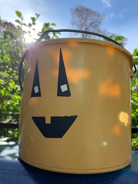 Mustard-yellow small cylinder bucket with wire handle, jack-o-lantern face adhered in black tape
