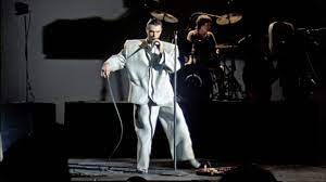 selects from Talking Heads' concert film Stop Making Sense. images of Byrne holding a microphone to camera, raising his arms, dancing in a big suit an with a lamp and full shots of the band.