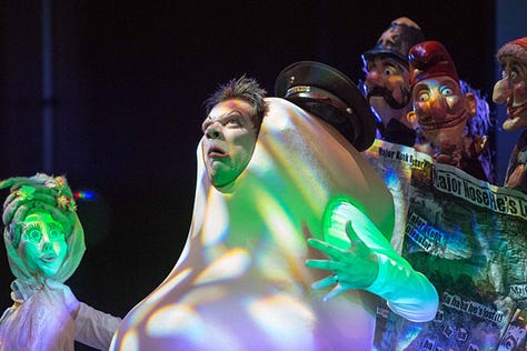 Image 1: "A dynamic stage performance featuring a person in a large, abstract costume resembling a nose, surrounded by puppeteers manipulating expressive characters. The scene is illuminated with colorful, dramatic lighting."  Image 2: "A performer dressed as a large nose interacts with the audience, holding a puppet of a woman with intricate details, while other puppets and props fill the vibrant stage, creating a surreal and theatrical atmosphere."  Image 3: "Close-up of a performer in a whimsical nose costume, exuding a dramatic expression, flanked by colorful puppets with exaggerated features. The scene is highlighted with vibrant stage lights."