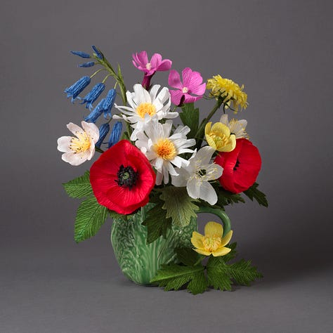 A beautiful arrangement of wildflowers in bright shades of pink, yellow, red, blue and white, presented in a green cabbage leaf mug, against a dark grey background. Two pictures of the author trying not to laugh, or breathe too hard, on the arrangements as she makes the final tweaks!