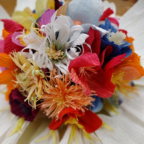 A giant paper daisy, created by artist Ling Warlow and local school children. White outer ray florets encircle a brightly coloured composite centre with the disc florets in an array of colours and flower shapes