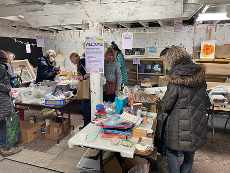 Swappers in a basement space that has filled up with tables and craft materials, clear jars with votes on having the swap again, and a Meadowlark Guide Co coffee mug from a friend who helped arrange materials.