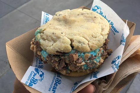 Three images of large delightful cookies with blue ice cream in the middle and toffee chunks or sprinkles around the outside edges of the ice cream, all being held in wrappers that say The Baked Bear.