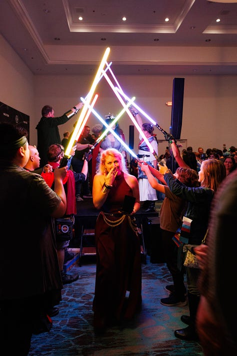 1: Cass on stage with the Halcy-Con event team, all arm-in-arm | 2: A massive circle of people hugging and swaying to a song; Cass is visible in the foreground, definitely trying to hide the fact that she's weeping by tucking her head in a friend's shoulder | 3: Cass beneath an arch of lightsabers, hand held to her mouth because there's really no hiding that she's crying this time.