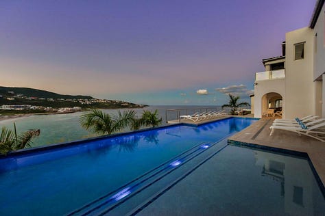 An aerial view of Villa Amalia perched on the cliffs of Oyster Pond, St. Martin, showcasing its sleek modern design with an expansive infinity pool overlooking the turquoise Caribbean Sea. The villa’s open-concept layout, floor-to-ceiling glass walls, and spacious outdoor terrace offer a perfect blend of indoor and outdoor luxury living, with lush greenery surrounding the property and the ocean visible in the distance.