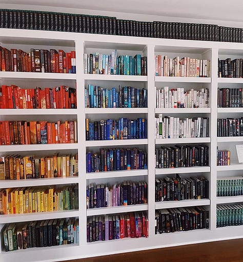 from left to right: a black lounge chair set on a dark hardwood floor, a white bookshelf filled with books arranged by color in rainbow order; a white freestanding bathtub set against a window