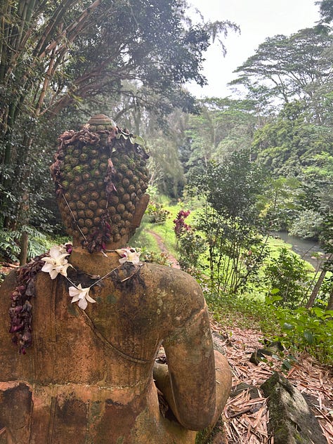 A white stick with Hawaiian on one side and English on another with the message "May Peace Prevail on Earth". The image in the center is of a stone buddha statue overlooking a ravine of tropical plants.