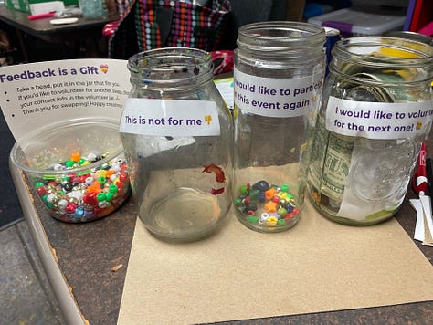 Swappers in a basement space that has filled up with tables and craft materials, clear jars with votes on having the swap again, and a Meadowlark Guide Co coffee mug from a friend who helped arrange materials.