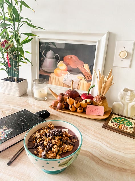 Close-up image of sand along a seashore and multi-hued sunset sky in the distance; image of an açaí breakfast bowl, journal, a framed painting, a bamboo plant, decorative wooden fruit, a vase, and a coaster on a kitchen dining table; image of tall beach grasslands in the foreground with buildings in the background backlit by the sunset light; dappled light through leaves of a mango tree and strung Tibetan prayer flags from the second-floor entrance to a yoga studio; image of a salad from a patio table and dining area surrounded by lush vegetation; image of two feet in sneakers on the sand with an infinity symbol drawn in the sand.