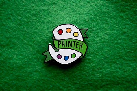 Three enamel pins with the words "WRITER," "PAINTER," and "ARTIST" emblazoned on the front.