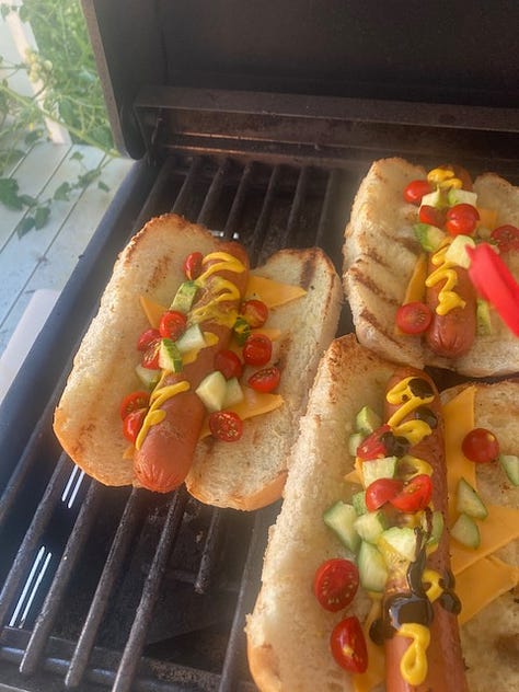 Grilled hotdogs being prepared on an outdoor grill with a number of ingredients and the buns.