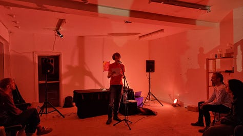 Three images of Stu reading in a room lit by red lights. Location: The Auxiliary Project, Middlesborough. July 2023