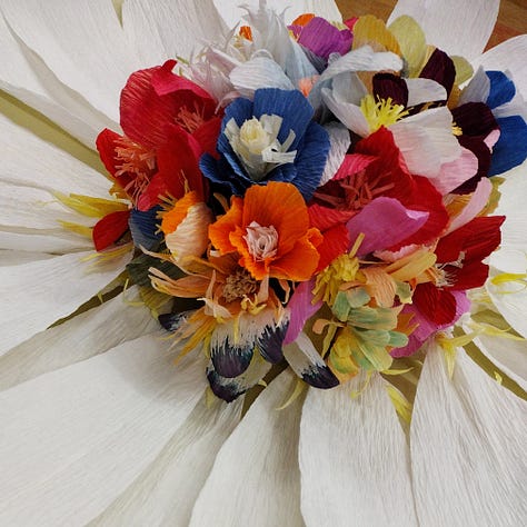 A giant paper daisy, created by artist Ling Warlow and local school children. White outer ray florets encircle a brightly coloured composite centre with the disc florets in an array of colours and flower shapes