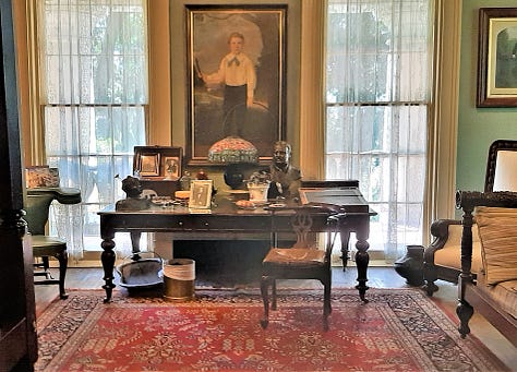 The interior of Houmas House is stately and beautiful with a wide center hall with large portraits on the wall. The second photo shows a fireplace and artwork; the third photo shows a desk in a stately room with a portrait in an ornate frame. The last photos are of bedrooms and a curving staircase.