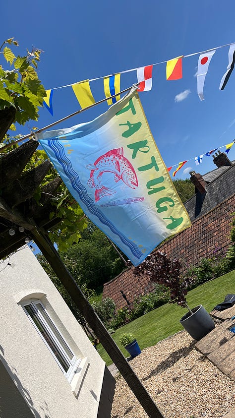 A blue and yellow flag with the boat's name "Tartuga" on it, with imagers of a fish and a pen (our dual passions) billowing against blue sky