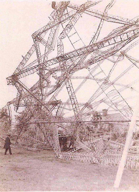 The zeppelin's metal scaffold structure lays in a field, several stories high. Officers inspect the structure and a crowd waits behind a taped cordon.