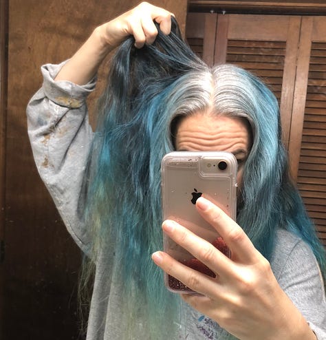 The author getting ready to re-dye and re-bleach her hair in sea-blues, pulls up the back of her hair where it's still brunette, in contrast to the silver roots on the top and sides of her head.