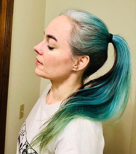 The author getting ready to re-dye and re-bleach her hair in sea-blues, pulls up the back of her hair where it's still brunette, in contrast to the silver roots on the top and sides of her head.