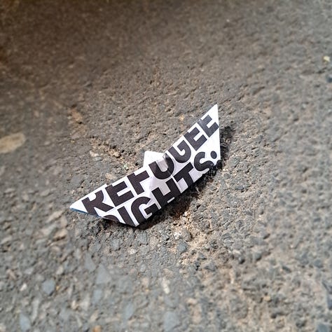 an origame paper boat left on the path. paper had 'refugee rights' written in block print across it.