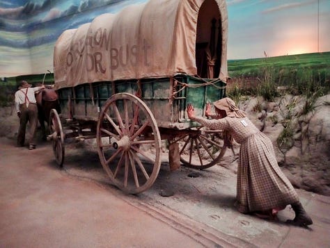 Three views of same tableau: Overview, showing a life-sized covered wagon being pulled by oxen and pushed from behind by woman in dress, as it heads toward the spire of Chumney Rock, a major landmark, and the sun sets. Close up of woman pushing, showing Oregon or Bust painted in large letters on side of covered wagon; view from front, showing man in hat cracking whip over four oxen pulling wagon