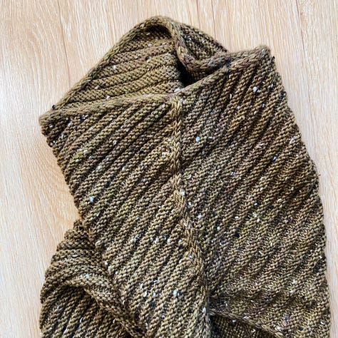 A brownish-green knitted cowl laying on a light brown floor in different positions.