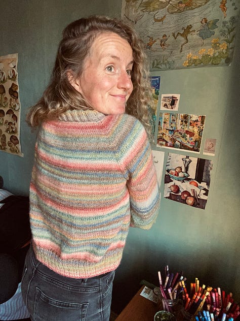 JB wearing a pastel coloured striped jumper that she knitted. She is standing in said jumper looking very pleased, with her thumbs up and a smile on her face.