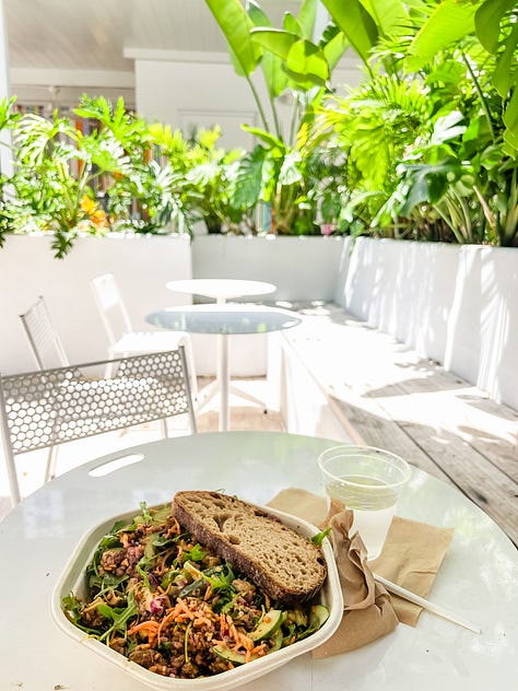 Close-up image of sand along a seashore and multi-hued sunset sky in the distance; image of an açaí breakfast bowl, journal, a framed painting, a bamboo plant, decorative wooden fruit, a vase, and a coaster on a kitchen dining table; image of tall beach grasslands in the foreground with buildings in the background backlit by the sunset light; dappled light through leaves of a mango tree and strung Tibetan prayer flags from the second-floor entrance to a yoga studio; image of a salad from a patio table and dining area surrounded by lush vegetation; image of two feet in sneakers on the sand with an infinity symbol drawn in the sand.