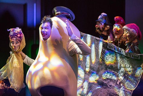 Image 1: "A dynamic stage performance featuring a person in a large, abstract costume resembling a nose, surrounded by puppeteers manipulating expressive characters. The scene is illuminated with colorful, dramatic lighting."  Image 2: "A performer dressed as a large nose interacts with the audience, holding a puppet of a woman with intricate details, while other puppets and props fill the vibrant stage, creating a surreal and theatrical atmosphere."  Image 3: "Close-up of a performer in a whimsical nose costume, exuding a dramatic expression, flanked by colorful puppets with exaggerated features. The scene is highlighted with vibrant stage lights."