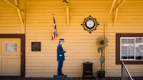 Three photos of Colfax. The first is the exterior of the local historical society building, an old wooden building painted yellow with large eaves and simple wooden brackets holding up the eaves. A door on the left has a hand written sign, "SHUT". From left to right is a bronze historical marker, a flag, an artistic lifesize cutout of a train conductor looking at his watch, an old clock with iron filligree, a wood burning stove, and a life-sized artist sculpture of a sunflower in a pot. The second photo are the rusty iron wheels and apparatus of an old rail car, including the armature for applying the brakes and a small section of rail to sit upon. Third photo is another exterior shot of the historical society, a closeup of objects outside the yellow building, including a primitive hand-painted sign of an old steam train on the tracks with mountains in the background set within a roundel around which the words, "COLFAX AREA HISTORICAL SOCIETEY" are painted. In front are some rusty milk barrels. A large round sign (bars of blue, white, and red with the words, "COLFAX CAN DO IT!" painted on it) sit on two of the barrels. A smaller sticker "TRUE" is affixed at the bottom of the sign.