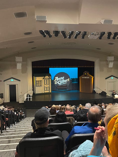 1) Caitlin, a white woman with dark brown hair, wearing a multicoloured dress with puffed sleeves standing in front of a large poster for Anne of Green Gables the Musical 2) The auditorium of a theatre with the crowd in their seats and the stage has a large screen with a green pattern and a silhouette of Anne of Green Gables. 3) A different auditiorium with the crowd in their seats, on stage you can see to building front set pieces and circular screen with 'Anne & Gilbert' written on it.