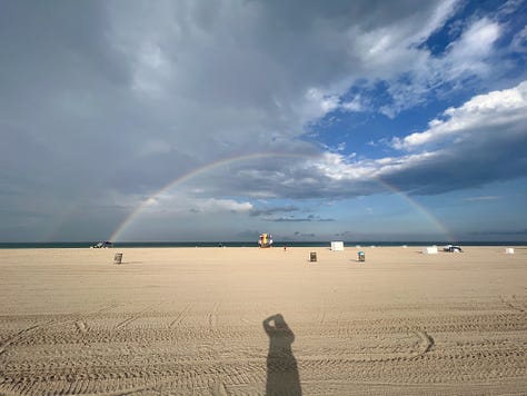 selfie photo of woman smiling in her apartment; selfie photo of three friends under a beach umbrella; photo of a beagle-King Charles Cavalier Spaniel mix dog looking down at the floor, taken from dog's eye level; photo of the photographer's legs and bare feet standing on thick, green blades of grass; photo of a microphone and computer screen with recording software; photo of a black and white cat lounging on a paved path of lush garden in the backyard of a building; photo of sunset at the beach with rays of pink and white and blue light over the horizon above the water; photo of cars driving in heavy traffic on a highway with billboards and buildings in the distance and white and gray clouds in the foreground and a clear blue sky in the background; wide-angle photo of a wide tan beach with the ocean in the far distance and a blue sky with white and gray swirling clouds above and a wide double rainbow over the shoreline.