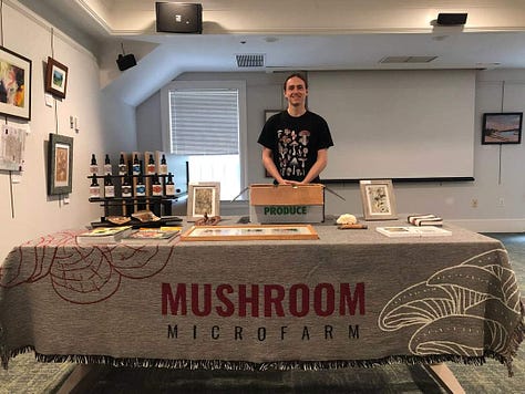 In the first photo James stands behind a table with a blanket printed witht he words "Mushroom Microfarm". Second picture, James and table are in the background with class attendees at forefront. Third picture, close up of tinctures on the table.