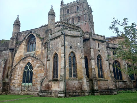 Exterior side of stone building with clock tower, interior with large stained glass window, interior with smaller stanined glass windows, altar, and ceiling decoration that looks like large star, large star, gift shop fll of merch in side chapel, cake slice and cup of tea