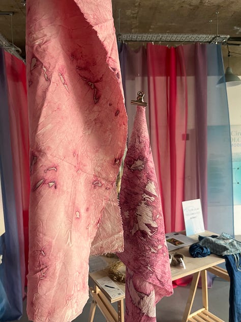 1-2. Exhibition table for 'pinks' from Ruth Lloyd and Colorifix showing fabric samples in petri dishes dyed directly with bacteria, and a vial of bacterial culture; 3. Pink bacteria-dyed fabrics hung up in the exhibition space with other brightly-coloured fabrics in the background.