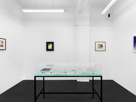 1) The table full of works is in the center of the image. It has a black frame and a clear top. There are also several frames of Sadao's work hanging on all the surrounding walls. 2) A straight down view of a table is seen showing many different drawings from Sadao. There are various sketches, and a cover of a gay magazine. 3) A clear showcase table is in the middle of a room, with a window to the left. There is a long horizontal frame on the wall full of Sadao's work on the back wall, and another black framed image on the right wall. The table in the center is full of his drawings.